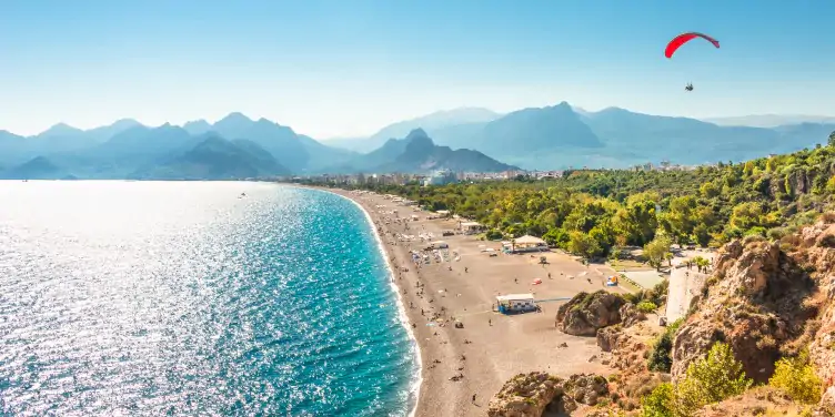 Birds eye view of a beach in Antalya, with tourists and locals enjoying the bright blue sea and sunny weather. With the cloudy mountains and a paraglider in the distance. 
