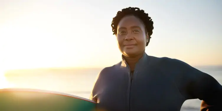 Lady standing on a beach with a surfboard with the sea in the distance. 