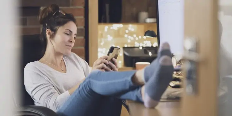 Middle aged woman sits with her feet on a desk looking at her phone