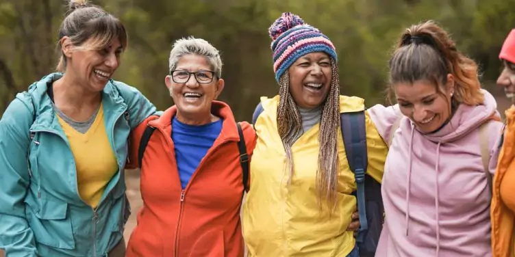 A group of five female friends arm in arm dressed in bright outdoor clothing ready to go hiking. 