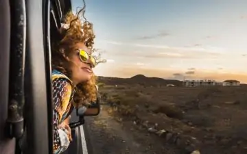 Young woman in sunglasses leans out of the car window while travelling on a solo trip