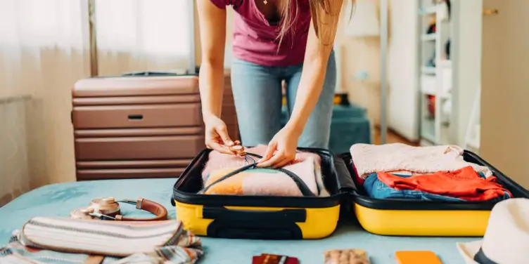 A young woman packs her brightly coloured suitcase surrounded by her possessions.