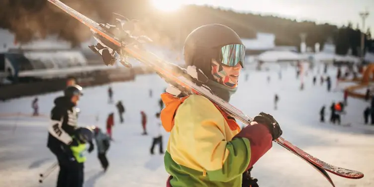 Skier wearing a helmet carries skis over her shoulder as she makes her way through a crowd of people at a popular skiing resort in Europe.