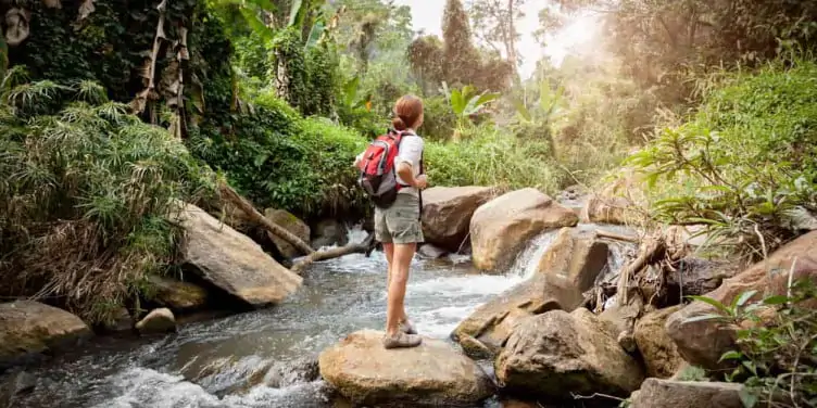 A young woman stands on a rock in the middle of a flowing river in the rainforest. 