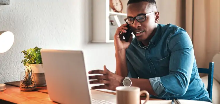 Man sat at desk using laptop while on the phone to his travel insurance company