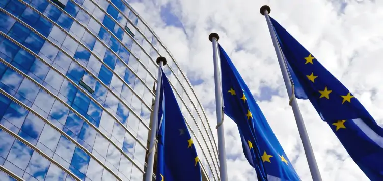 EU flags blowing in the wind next to a tall glass building with blue skys surrounding it. 