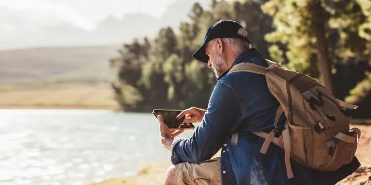 Man sitting on a rock, looking at his tablet, surrounded by a beautiful lake. 