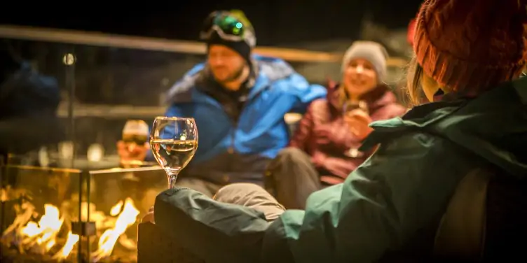 Group of skiers enjoying après-ski around a warm fire, with a glass of wine.
